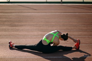 Güzel atletik bayan stadyumda esneme hareketleri yapıyor, esneklik antrenmanı yapıyor, atletik kadın bacak aralarına oturuyor ve elini bacağına uzatıyor, sağlıklı bir yaşam tarzı ve günlük egzersiz konsepti öneriyor.