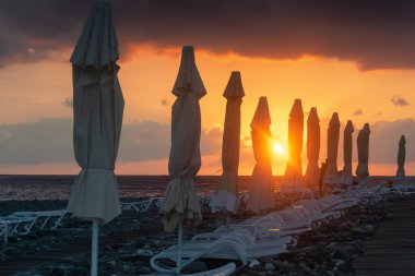 Sahilde gün batımı. Güneş gökyüzünü kırmızı ve turuncuya boyadı. Güneş ışınları geceleri kapalı olan plaj şemsiyelerine dokunuyor. Günbatımının parlak renkleriyle gece kumsalı