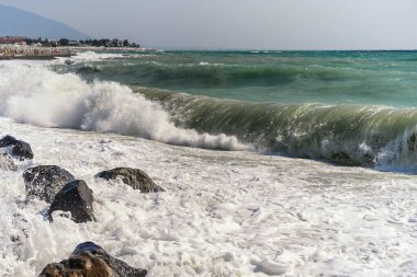 Sahilde dalgalar. Rüzgarlı hava, sahildeki birçok dalga deniz manzarasını canlandırıyor. Ön planda çakıl taşları üzerinde koyu taşlar, turistler uzakta dinleniyor. Dağların sislerinde
