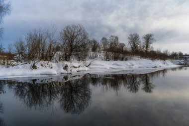 Nehirli kış manzarası. Beyaz kar kıyılarda uzanıyor. Ağaçlar suya yansıyor. Mavi gökyüzü ve beyaz bulutlar. Şiddetli dondan kaynaklanan hafif bir sis. kış günü