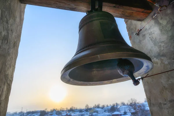 a large bell on the bell tower. cast iron bell with curls and tinting for metal. brick walls with openings. the winter city is visible below. sunny winter day on a tall ancient tower