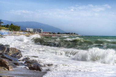 Kumsal ve fırtınalı deniz. Güçlü dalgalar kıyıdaki büyük karanlık taşlara vuruyor. İnsanlar kumsalda dinleniyor ve uzaklarda mavi bir sis içinde dağlar var.. 