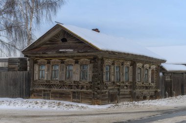 Güneşli bir kış gününde oyulmuş mimari unsurları olan eski ahşap bir ev. Çok güzel pencereler, tahta kaldırım, evin yanında uzun bir ağaç ve mavi bir gökyüzü var.. 