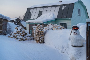Kar yığınları, kardan adam ve kış için yakacak odunları olan kırsal bir ev. Fotoğraf, Kuzey Urallar 'da (Rusya) bulutlu bir kış gününde çekildi. Atalarımızın gelenekleri, özen ve rahatlama 