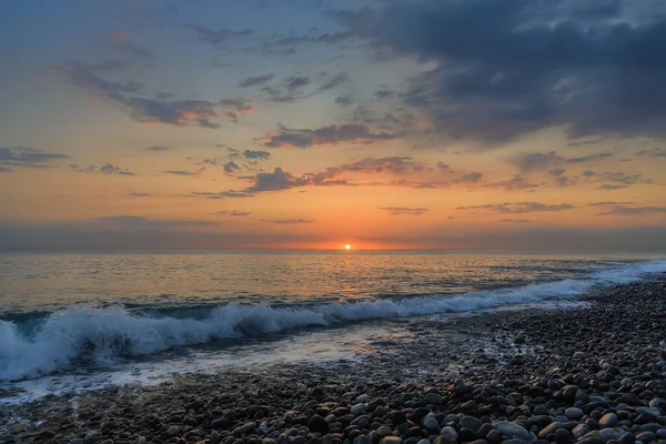Sunset over the sea. In the foreground, a wave running onto a pebbly shore with white foam. The dark blue sky with clouds, on the horizon line the orange colors of the sunset and the circle of the sun. reflection in water