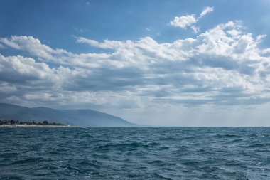Önünde dalgalar, uzağında dağlar olan deniz manzarası. Güzel mavi bir gökyüzü. Uzakta garip beyaz bulutlar yüzüyor. Sahilde küçük bir kıyı kenarı görünür.