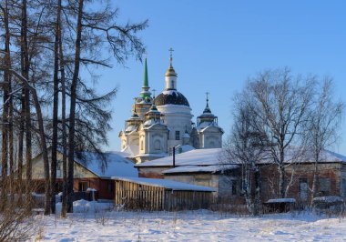 Güneşli bir kış gününde kubbeleri ve çan kulesi olan eski bir kırsal kilise manzarası. Uzun ağaçlar ve beyaz kar. Eski köy evleri görünür. Byngi Köyü (Sverdlovsk bölgesi, Rusya) 