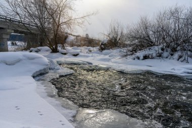 Nehirli bir manzara. Buz gibi bir kış gününde çözülmemiş, hızlı akan bir nehir. Gün ortası güneşi buzlu kıyılardan yansıyor. Çalılar ve ağaçlar sahneyi çerçeveliyor. Ural (Rusya) 