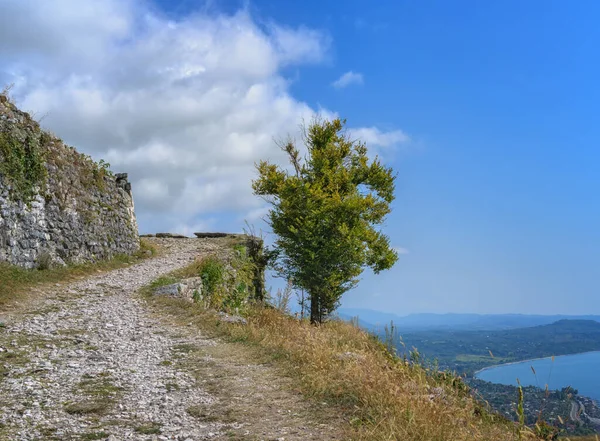 Rocky path in the mountains along the walls of the old fortress on a summer day. An old lonely tree is soloing against a blue sky with textured white clouds. Below the mountain is a blue sea with a beach.