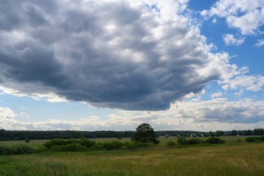Koyu gri bulutlu yaz manzarası. Kalıplı bir bulut, beyaz-mavi gökyüzüne karşı sololar yapıyor. Doğa, yaklaşan bir fırtınayı bekliyor. Uzakta bir köy görünüyor.. 