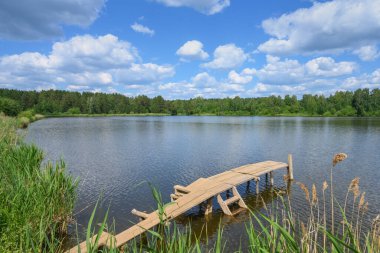 Ağaçlarla çevrili bir orman gölü ve üzerinde kümülüs beyaz bulutlar yüzen mavi bir gökyüzü olan yaz manzarası. Balıkçılık ve yüzme için tahta geçitler dikkat çeker. Rusya, Ural 