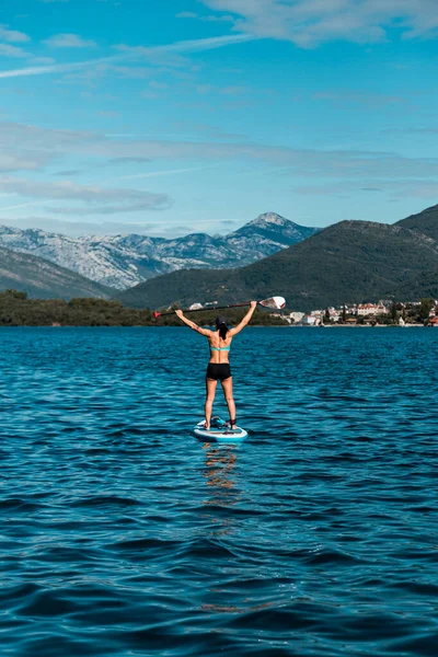 Young athletic woman stands on a SUP or stand up paddle board board in the Mediterranean with a beautiful view to the mountains