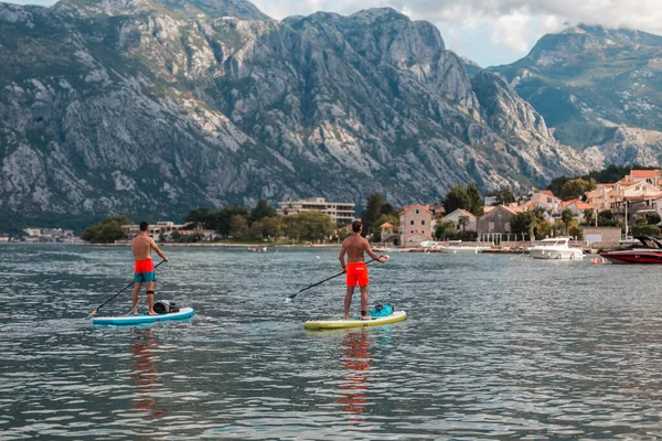Karadağ 'ın güzel bir köyüne bakan Kotor Körfezi' nde iki atletik adam bir SUP 'nin üzerinde ayakta duruyor.
