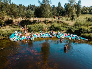 Bir sürü renkli SUP, güneşli bir yaz gününde nehir kenarındaki sörf tahtalarını kaldırır. İnsansız hava aracından görüntüle