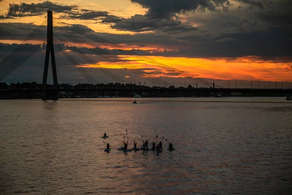 Group of people enjoy paddling with SUP stand up paddle boards in the river at sunset. View of Vansu bridge in the background. Active summer vacations with paddle board.