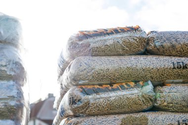 Bags of wood pellets wrapped in clear plastic are stacked outdoors under sunlight. Cylindrical pellets are visible through the transparent packaging.