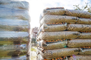 Rows of 15kg wood pellet bags wrapped in clear plastic sit outdoors under sunlight, with faint houses and trees visible in the distance.