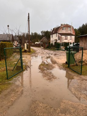 Su birikintileri olan çamurlu bir toprak yol yeşil metal bir kapıdan geçerek kırmızı çatılı, ağaçlar ve elektrik direkleriyle çevrili iki katlı bir eve doğru gidiyor..