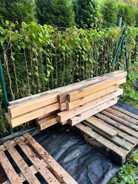 Wooden beams rest on pallets over a black tarp with a grid pattern. A green wire fence with climbing plants and tall hedges frame the scene.