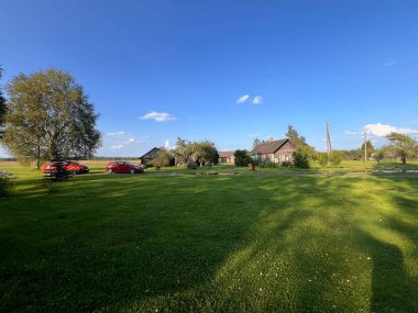 Ağaçlarla ve ahır gibi ahşap binalarla çevrili çimenlik bir arazide kırmızı arabalar ve mavi gökyüzünün altında elektrik hatları var..