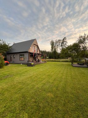 Dik çatılı modern gri bir ev ağaçlarla çevrili, bakımlı bir çimenlikte oturmaktadır. Sol tarafta kısmen görünen kırmızı bir araç var..