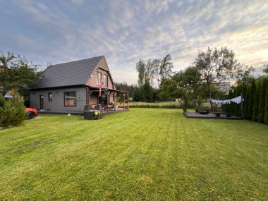 Koyu bir çatısı, verandası, verandası ve çamaşır ipi olan modern gri bir ev. Ağaçlar ve çalılar, parçalı bulutlu bir gökyüzünün altındaki çimlerin sınırında..