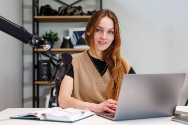 Genç bir kadın bir masada dizüstü bilgisayarı, defteri ve mikrofonuyla kayıt yapıyor. Metal rafta kamera, bitki ve ses ekipmanları var. Yumuşak ışık ve sığ alan derinliği.
