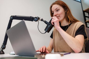 Genç bir kadın, modern bir ofisin düzenli bir masasında, parlak ışık ve sığ alan odaklama detaylarının derinliği olan bir masada dizüstü bilgisayar kullanırken, mikrofondaki siyah yoğunlaştırıcı mikrofona konuşur..