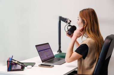 Bir kadın, küçük bir stüdyoda dizüstü bilgisayarı, pop filtresi ve patlama koluyla askıya alınmış bir kardiyo mikrofonuna kayıt yapıyor. Yumuşak ışıklandırma bile onun orta oturum odağı vurgular.