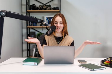 Genç bir kadın beyaz bir masada kayıt yapıyor, mikrofona konuşuyor, elleri havada, dizüstü bilgisayar, kulaklık, notlar, fotoğraf makineleri ve kitaplardan oluşan raflar, hafif bir ışık bile var..