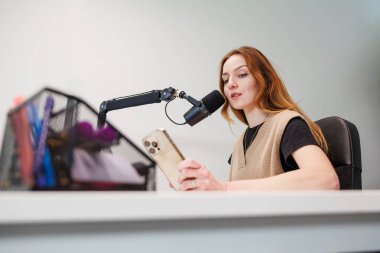 Genç bir kadın mikrofona doğru konuşur ve stüdyo masasındaki akıllı telefondan okur. Yumuşak ışık ve sığ alan derinliği modern içerik oluşturmaya odaklanıyor.