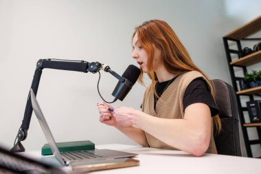 Genç bir kadın mikrofona doğru konuşur, bir dizüstü bilgisayarı kontrol eder ve minimalist bir ev stüdyosunda hafif ışık ve sığ bir alan derinliğinde küçük bir ses cihazı tutar..