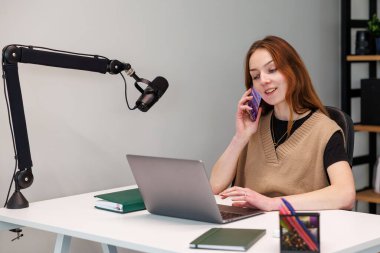 Beyaz bir masada oturan, akıllı bir telefonla konuşan ve dizüstü bilgisayarda çalışan bir kadın. Eklemli bir patlama kolu ve yoğunlaştırıcı mikrofon modern bir ev stüdyosunda solunda oturuyor..
