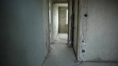 Kablolu, borulu ve pencereli tamamlanmamış apartman koridoru. Korunmasız kablolar, çıplak alçı, beton zemin, toz ve serin ışık. Sığ alan derinliği kaba bir doku gösteriyor.