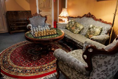 Süslü bir oturma odası yuvarlak bir masanın etrafına oyulmuş koltuklar sunar gümüş ve altından yapılmış cam bir satranç tahtası, sıcak bir lamba ışığı ve İran tarzı bir halı..