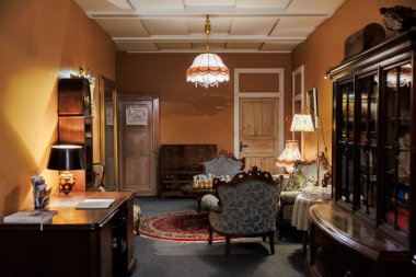 Antika dolaplar, yazı masası ve İran halısı etrafında Victoria dönemi oturma düzeni. Altın lamba, toprak duvarları, parlak odunları ve döşemeleri ısıtır..