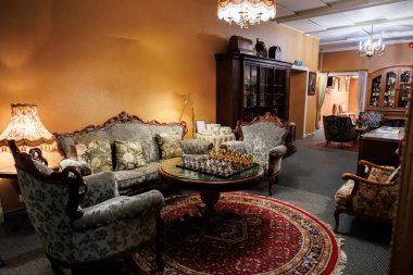 Viktorya dönemi oturma odasında maun koltuklar, cam ve metal satranç seti, kırmızı İran tarzı halı, avizeler, lambalar, dolaplar, çerçevelenmiş sanat eserleri ve koridor bulunur..