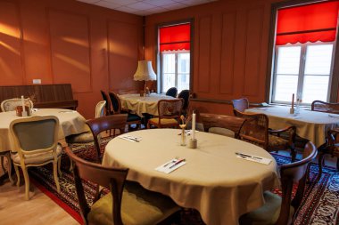 Konforlu bir yemek odasında bej kumaşlar, klasik sandalyeler, desenli halılar ve kırmızı jaluzili masalar var. Bir zemin lambası ve küçük dik bir piyano teras duvarlarının yanında oturur..