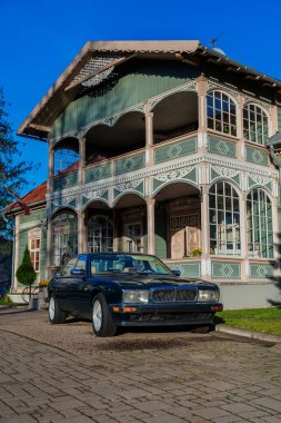 Koyu renkli bir Jaguar sedanı, iki katlı ahşap bir villada, zencefilli ekmek budanmış, kemerli verandalı, elmas kaplamalı ve parlak gün ışığında pastel yeşil paneller üzerinde duruyor..