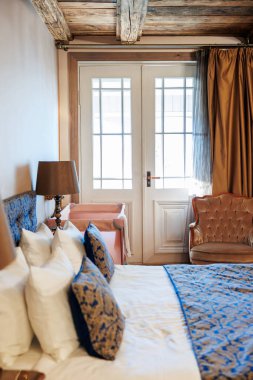 Mavi tekstil desenli çift kişilik bir yatak pirinç bir lambanın, kadife koltuğun ve pembe bankın yanında oturur. Gün ışığı altın perdelerin arasından geçerken Fransız kapılarının önünde..
