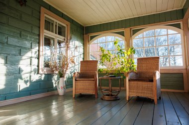 İki hasır koltuk ve bir yuvarlak masa kapalı bir verandada döşeme üzerinde oturur. Kapalı duvarlar ve kemerli pencereler sonbaharın sonlarında gevrek gölgeler bırakır..