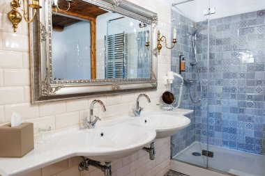 Kavisli beyaz leğenleri olan çift kibir gümüş bir aynanın altında ve pirinç şamdanların yanında gri fayanslar, krom musluklar ve bir havlu radyatörü olan cam bir duşun yanında oturur..