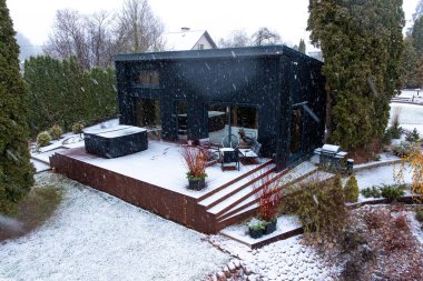 Kömür kömürü, siyah modern bir ev yükseltilmiş bir güvertede, buzlu sandalyeler ve kare bir jakuzi, hafif kar yağışı, yüksek yemyeşil bir sınır, banliyöde gün ışığı soluyor..