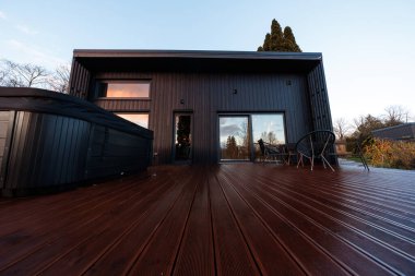 Düşük açı tek katlı bir ev, dikey metal kaplama, geniş cam kapılar, kırmızı kahverengi kompozit bir güverte, iki sandalye, masa ve alacakaranlıkta koyu renkli bir jakuzi gösteriyor..