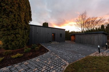 Minimalist siyah kereste evi ve ayrık garaj kaldırım taşları ve karanlık kaldırımların yanında oturur, bir sıcak giriş ışığı açık, kozalaklı ağaçlar ve çıplak ağaçlar mevcut, yumuşak altın saat ışığı.