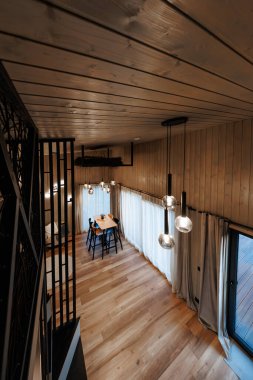 Üst merdiven manzarası ahşap kaplı çatı katı, çift katlı oda, geniş tahta zemin, düz duvarlar, katlı perdeler, masadaki siyah sandalyeler, küre kolyeler, cam küme, gündüz ışığı.