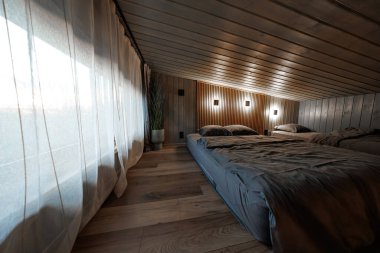 Gri çarşaflı üç katlı yataklar eğimli bir ahşap tavanın altında, kare şamdanlar slat duvarda parlıyor, perdeler yumuşuyor gün ışığı, bitki ve ahşap döşeme dokusu ekleniyor.