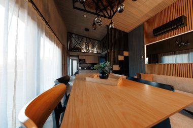 Geniş açı, ağaç masa, minimalist sandalyeler, perdeler, kompakt mutfak, asma kat korkulukları, kolye küre ışıkları, slat paneller ve havalandırma üniteleri gösteriyor..