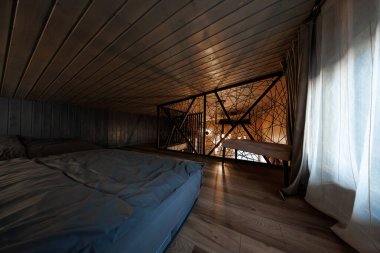 Alçak tavanlı bir çatı katında, perdelerin yanında tahta döşeli bir döşek var. Sıcak kolye ışıkları, akşam ışığında çapraz kablolarla siyah metal bir tırabzanın altında parlıyor..