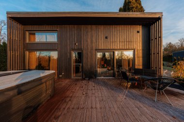 Dikdörtgen bir kulübe dikey kömürleşmiş ahşap, eğimli duvarlar ve düşük eğimli çatı geniş bir güverteye açılıyor siyah sandalyeler, bank, masa ve sıcak küvet ile geç gün ışığı.